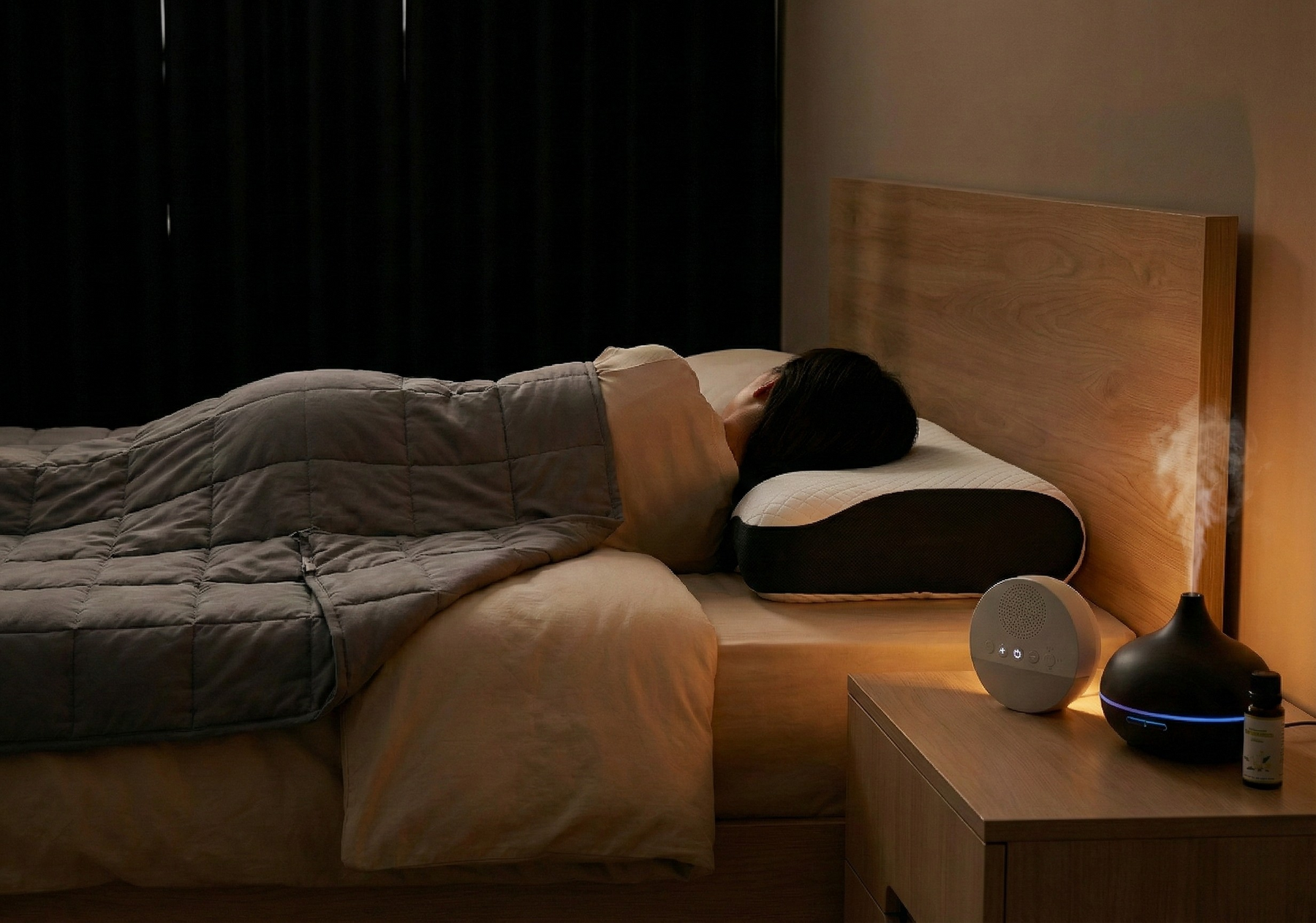 Personne allongée dans une chambre apaisée avec oreiller ergonomique, couverture lestée, machine à bruit blanc et diffuseur d’huiles essentielles, illustrant un environnement pensé pour le repos.